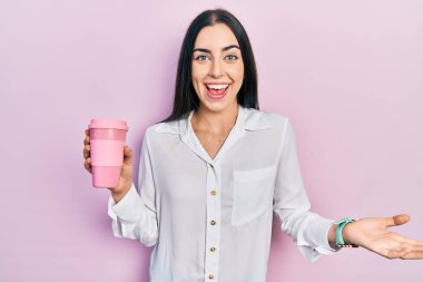 Mavi gözlü güzel bir kadın bir fincan kahve içerek başarıyı kutluyor mutlu bir gülümsemeyle ve elini kaldırarak kazanan bir ifadeyle. 