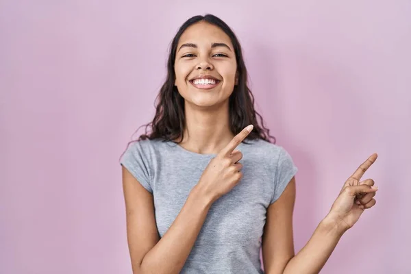 年轻的巴西女人穿着宽松的T恤 背景粉红 面带微笑 看着摄像机 用双手和手指指向旁边 — 图库照片