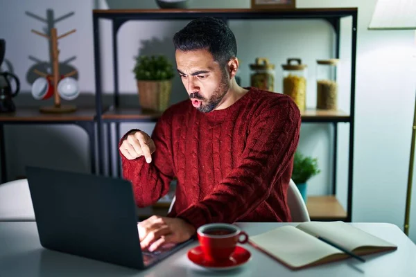 Young hispanic man with beard using computer laptop at night at home pointing down with fingers showing advertisement, surprised face and open mouth