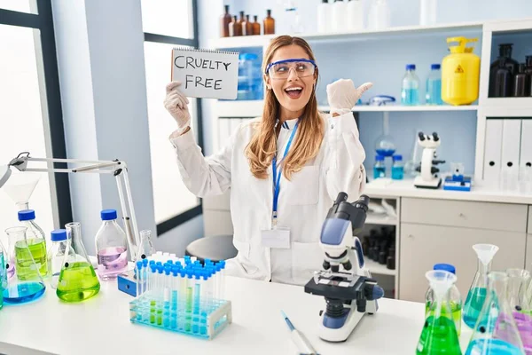 Young blonde scientist woman working on cruelty free laboratory pointing thumb up to the side smiling happy with open mouth