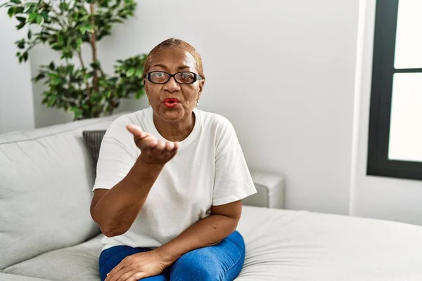 Mature hispanic woman sitting on the sofa at home looking at the camera blowing a kiss with hand on air being lovely and sexy. love expression.