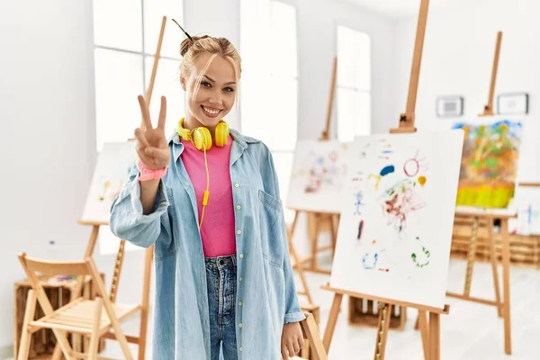 Young caucasian girl at art studio showing and pointing up with fingers number two while smiling confident and happy. 