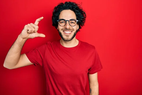 カジュアルなTシャツとメガネを身に着けているハンサムなヒスパニック系の男は 指を見て カメラで小さなサイズの看板を行う手で笑顔と自信を持ってジェスチャー 概念を測定し — ストック写真
