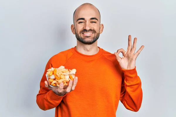 Young bald man holding potato chip doing ok sign with fingers, smiling friendly gesturing excellent symbol