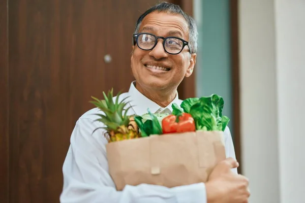 Middle age southeast asian man smiling holding a bag of fresh groceries standing by home door