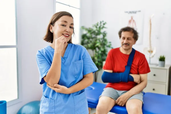 Middle age doctor woman with patient with arm injury at rehabilitation clinic smiling looking confident at the camera with crossed arms and hand on chin. thinking positive.