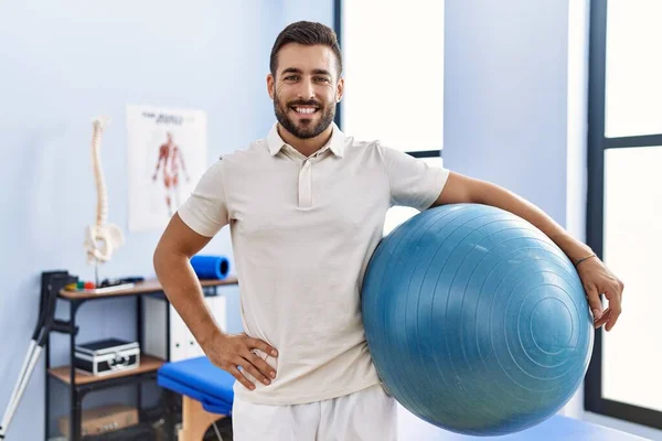 Przystojny Hiszpan Trzymający Pilates Ball Klinice Rehabilitacyjnej Wyglądający Pozytywnie Szczęśliwie — Zdjęcie stockowe