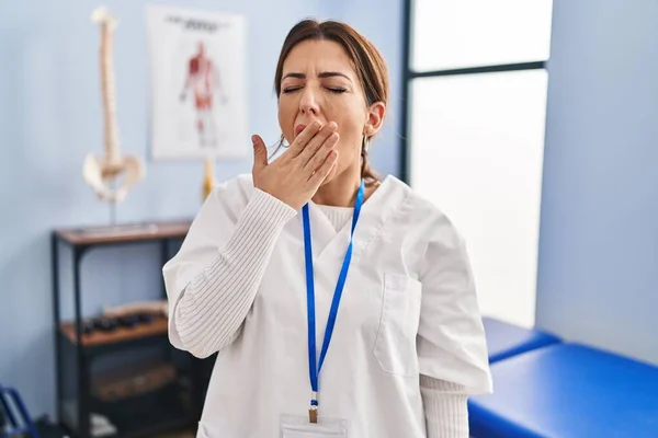 Young brunette woman working at pain recovery clinic bored yawning tired covering mouth with hand. restless and sleepiness.