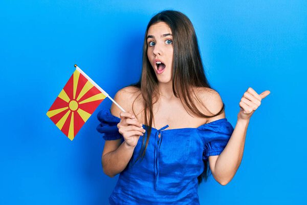 Young brunette teenager holding macedonian flag pointing thumb up to the side smiling happy with open mouth 