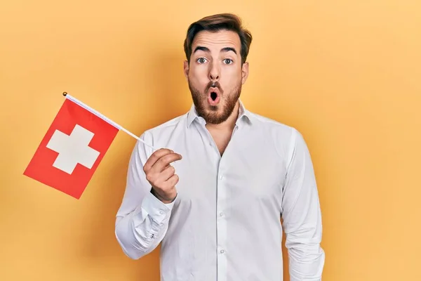 髭を生やしたハンサムな白人男性がスイス国旗を掲揚し 驚きのために口を開けて驚いて 不信の顔 — ストック写真