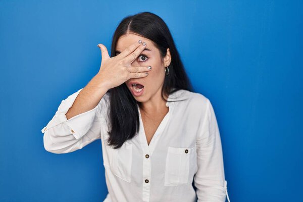 Молодая латиноамериканка, стоящая на синем фоне, заглядывая рукой в лицо и глаза, смотрит сквозь пальцы со смущенным выражением лица.. 