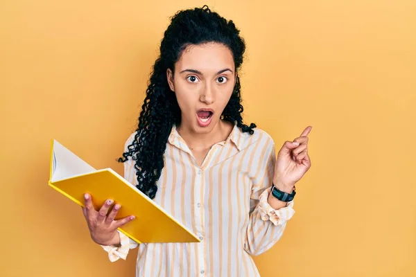 Young Hispanic Woman Curly Hair Holding Book Surprised Pointing Finger — Stockfoto