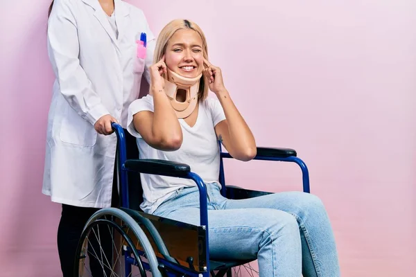 Beautiful blonde woman sitting on wheelchair with collar neck covering ears with fingers with annoyed expression for the noise of loud music. deaf concept.