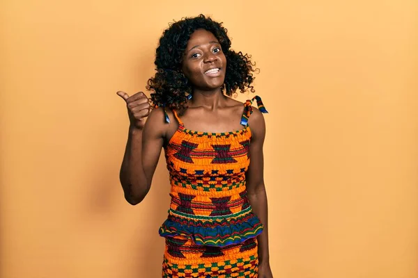 若いアフリカ系アメリカ人の女性は幸せな顔をして笑顔で伝統的なアフリカの服を着て親指を上げて側面を指差す — ストック写真