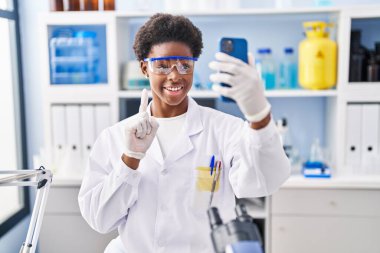 African american woman working at scientist laboratory doing video call with smartphone smiling with an idea or question pointing finger with happy face, number one 