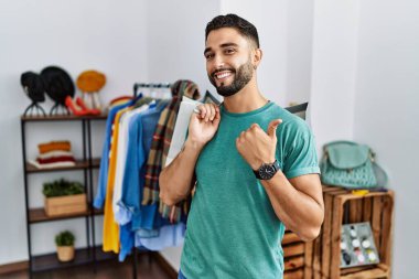 Perakende mağazasında sakallı, alışveriş torbaları tutan genç yakışıklı adam arka tarafı işaret ediyor, el ve başparmakları havada, kendinden emin gülümsüyor. 