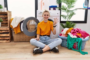 Young blonde woman doing laundry sitting by washing machine showing and pointing up with fingers number four while smiling confident and happy. 