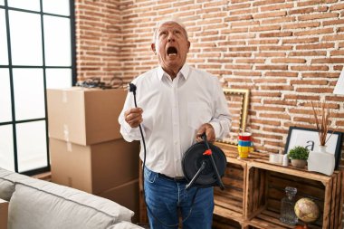 Senior man with grey hair holding extension plug angry and mad screaming frustrated and furious, shouting with anger looking up. 