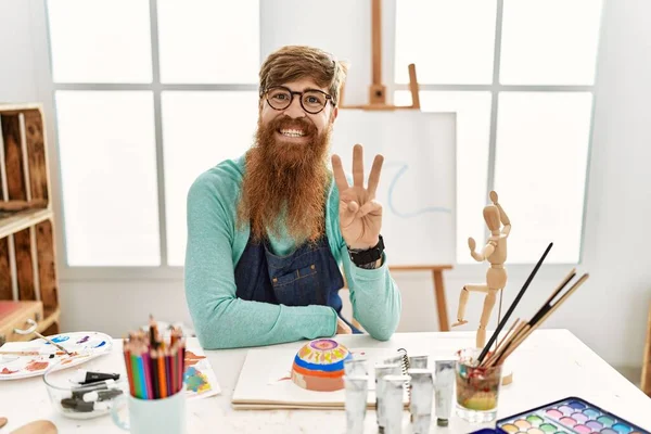 Redhead man with long beard painting clay bowl at art studio showing and pointing up with fingers number three while smiling confident and happy.