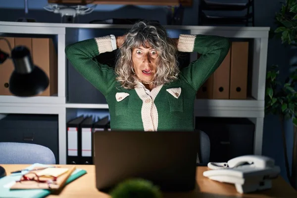 Middle age woman working at night using computer laptop crazy and scared with hands on head, afraid and surprised of shock with open mouth