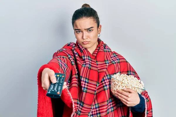 Young caucasian girl wearing blanket eating popcorn using tv control depressed and worry for distress, crying angry and afraid. sad expression.