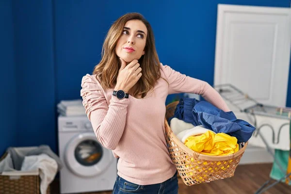 Young woman holding laundry basket with hand on chin thinking about question, pensive expression. smiling with thoughtful face. doubt concept.