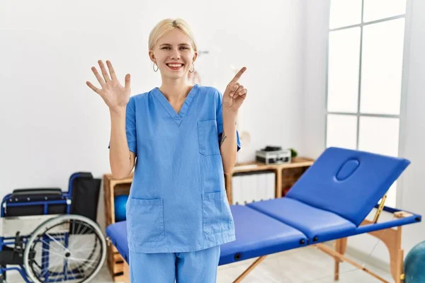 Beautiful caucasian physiotherapist woman working at pain recovery clinic showing and pointing up with fingers number six while smiling confident and happy.