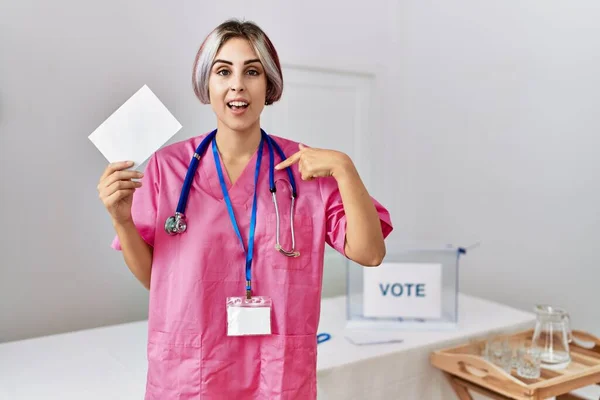 Young nurse woman at political campaign election holding envelope pointing finger to one self smiling happy and proud