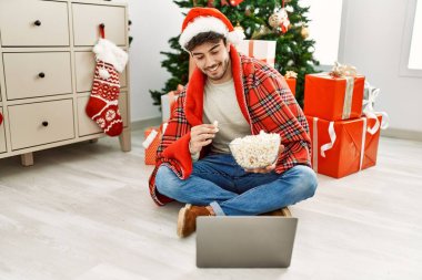 Young hispanic man wearing christmas hat watching movie and eating popcorn at home.