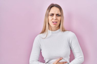 Pembe arka planda beyaz süveter giyen genç sarışın kadın midesi üstünde el ele çünkü hazımsızlık, acı verici bir hastalık kendini iyi hissetmiyor. Ağrı kavramı. 