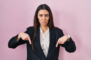 Pembe arka planda iş elbisesi giymiş genç esmer kadın üzgün ve üzgün görünüyor, parmaklarıyla yönü gösteriyor, mutsuz ve depresif.. 