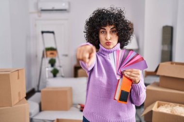 Yeni ev duvarının rengini seçen kıvırcık saçlı genç esmer kadın kamerayı ve seni işaret ediyor. Kendine güveni tam bir jest. 