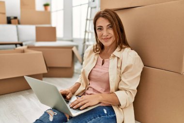 Orta yaşlı, beyaz bir kadın yeni evinde dizüstü bilgisayar kullanıyor..