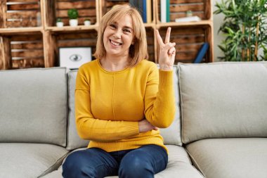 Middle age caucasian woman wearing casual clothes sitting on the sofa at home smiling with happy face winking at the camera doing victory sign. number two. 