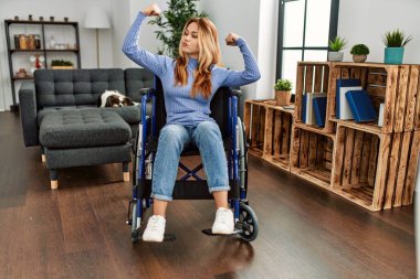 Young beautiful woman sitting on wheelchair at home showing arms muscles smiling proud. fitness concept. 