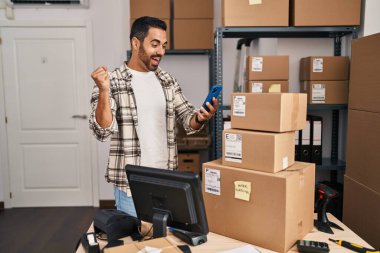 Sakallı genç İspanyol adam küçük işletme ecommerce çalışıyor. Akıllı telefonuyla gururla bağırıyor, zaferi ve başarıyı kutluyor. Kolunu kaldırarak heyecanlanıyor. 
