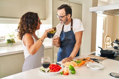 Orta yaşlı İspanyol çift mutfakta yemek pişiriyor ve bardaklara şarap döküyor.