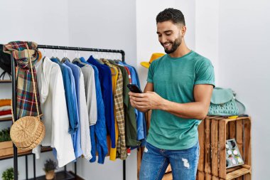 Genç Arap müşteri akıllı telefon kullanıyor giyim mağazasında elbise rafının yanında duruyor.