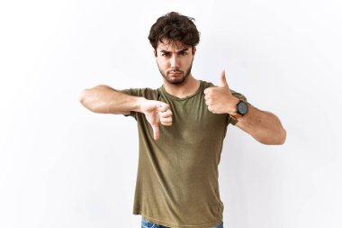 Hispanic man standing over isolated white background doing thumbs up and down, disagreement and agreement expression. crazy conflict 