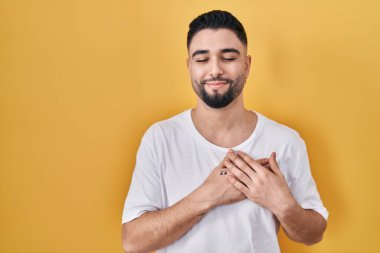 Sarı arka planda rahat bir tişört giyen genç yakışıklı elleri göğsünde, gözleri kapalı ve yüzünde minnet dolu bir jest ile gülümsüyor. sağlık kavramı. 