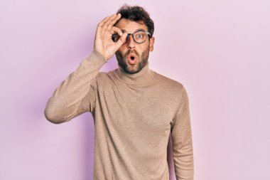 Balıkçı yaka kazak ve gözlük takan sakallı yakışıklı adam şaşırmış yüz ifadesiyle şaşırmış, parmaklarının arasından bakıyor. İnançsız ifade. 