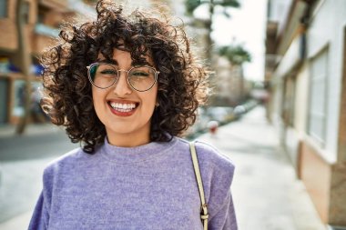 Orta Doğulu genç bir kadın gülümsüyor. Sokakta gözlük takıyor.