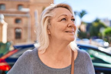 Orta yaşlı sarışın kadın sokakta kendinden emin bir şekilde gülümsüyor.