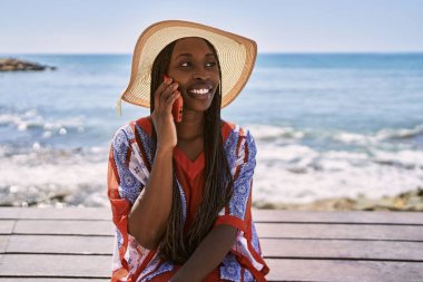 Genç Afrikalı Amerikalı turist, kumsalda bankta oturmuş akıllı telefondan konuşuyor..