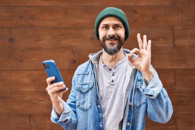 Young hispanic man using smartphone doing ok sign with fingers, smiling friendly gesturing excellent symbol 