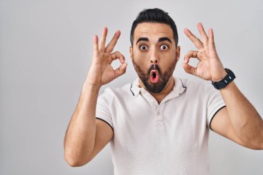 Beyaz arka planda sıradan kıyafetler giyen, sakallı genç İspanyol adam şaşırmış ve şaşırmış görünüyor. Tamam sembolünü parmaklarıyla yapıyor. çılgın ifade 