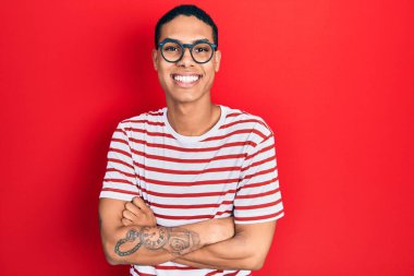 Genç Afro-Amerikalı adam günlük kıyafetler ve gözlük takıyor. Pozitif ve mutlu görünüyor. Kendine güvenen bir gülümsemeyle dişlerini gösteriyor. 