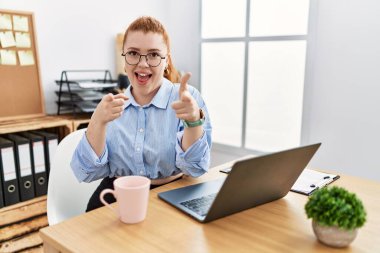 Ofiste çalışan genç kızıl saçlı kadın bilgisayarını kullanarak kamerayı mutlu ve komik yüzüyle işaret ediyor. iyi enerji ve titreşimler. 