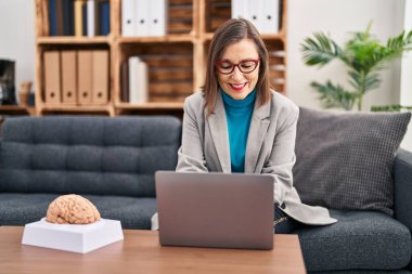 Middle age woman psychologist smiling confident using laptop at psychology center