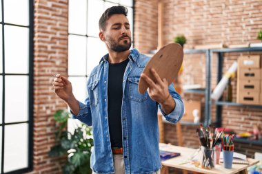 Resim stüdyosunda elinde fırça ve palet tutarak gülümseyen İspanyol genç adam.